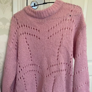 Rosa stickad tröja från Gina Tricot - Mysig rosa stickad tröja från Gina Tricot med ett dekorativt hålmönster. Perfekt för kyliga dagar med sin mjuka och bekväma passform. Tröjan har långa ärmar och en ribbad krage.