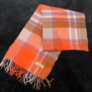 Rutig halsduk från Burberry - Säljer en snygg rutig halsduk från Burberry i orange, lila och beige toner. Den har fransar i ändarna och är perfekt för att hålla dig varm under kyliga dagar. Ett stilrent och klassiskt val för din garderob. 100% cashmere.