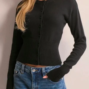 Snygg kofta från Nelly - Snygg svart kofta med knappar framtill och ribbad midja. Den är croppad och har långa ärmar, perfekt för en stilren look.