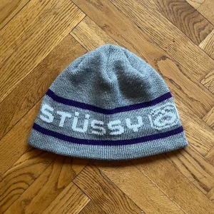 Stüssy Mössa - Vintage grå mössa från Stüssy med ett brett vitt och lila band runtom och Stüssy-loggan. Bra skick, 8/10. 
