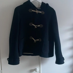 Marinblå duffeljacka från Aubin & Wills - Snygg svart duffeljacka från Aubin & Wills med klassiska träknappar och huva. Perfekt för kyliga dagar med sin stilrena design och praktiska fickor. Ett måste för garderoben! Har några noppror men det går enkelt att ta bort! De står stl 10 men tror de motsvarar Xs/S då jag bär den storleken och den passar perfekt! I äkta ull