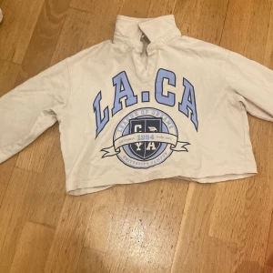 Beige tröja från H&M med LA-tryck - Säljer en beige tröja från H&M med stort blått LA-tryck och texten 'League of Dreams' samt 'California 1994'. Tröjan har en krage och är croppad i modellen. Perfekt för en avslappnad stil.