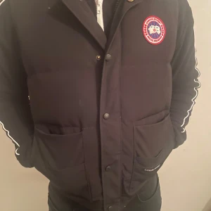 Svart pufferjacka från Canada Goose - Säljer en svart pufferjacka från Canada Goose med ett broderat märke på bröstet. Jackan har långa ärmar med vita detaljer längs sidorna och knappar framtill. Perfekt för kyliga dagar!