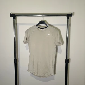 Beige t-shirt från Gymshark - Snygg beige t-shirt från Gymshark i storlek XS. Tillverkad av återvunnen polyester, bomull och elastan för en bekväm passform. Perfekt för träning med korta ärmar och stilren design med logga på ryggen.