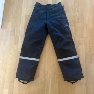 Svarta byxor från T. Sportswear - Svarta termobyxor från T. Sportswear med reflexdetaljer runt benen. Perfekta för utomhusaktiviteter med slitstarkt material.