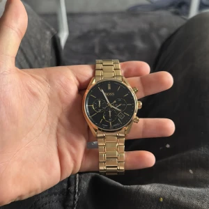 Guldfärgad klocka från BOSS - Snygg guldfärgad klocka från BOSS med svart urtavla och flera sub-dials. Armbandet är i metall och har en elegant design. Perfekt för att ge en lyxig touch till din stil. Helt nytt och kommer med tagen och plasten bakom urtavlan aldrig använt nypris 6200:- priset kan diskuteras vid snabb och smidig affär