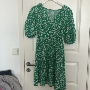 Grön blommig klänning - Charmig grön klänning med vitt blommönster. Tillverkad i 100% polyester, perfekt för vår och sommar. Klänningen har korta ärmar och en bekväm passform.
