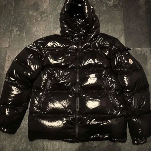 Moncler maya jacka glansig  - Snygg svart glansig pufferjacka med huva och dragkedja. Perfekt för kalla dagar med sin vadderade design. Jackan har långa ärmar och två fickor med dragkedja framtill.