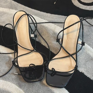 Svarta sandaler med snörning - Eleganta svarta sandaler med hög klack och smala remmar som snörs runt foten. De har en fyrkantig tå och en stilren design som passar perfekt för festliga tillfällen.