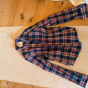 Rutig skjorta från Abercrombie & Fitch - Snygg rutig skjorta från Abercrombie & Fitch i blått, rött och gult. Skjortan har långa ärmar och knappar framtill. Perfekt för en avslappnad stil med en touch av färg.