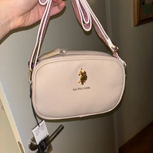 Beige axelväska från U.S. Polo Assn. - Snygg beige axelväska från U.S. Polo Assn. med ett stilrent märke i guld på framsidan. Väskan har en justerbar axelrem i vitt och rött samt en praktisk dragkedja upptill. Perfekt för att bära dina nödvändigheter med stil.