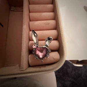 Silverfärgad ring med rosa hjärta och kaninöron - Unik ring i silverfärg med ett stort rosa hjärta i mitten och dekorativa kaninöron. Perfekt för att ge en lekfull touch till din stil. Ringen har även små stenar runt hjärtat för extra glans. Ringen är köpt av en smallbussnies här på Plick. DEN ÄR INTE SJUSTERBAR!!!!🌸🙏🏻