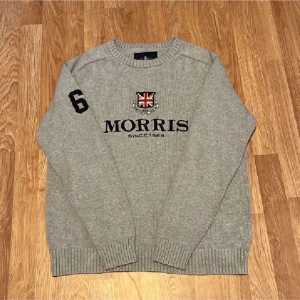 Grå stickad tröja från Morris - Säljer en stilren grå stickad tröja från Morris med broderad logga och nummerdetalj på ärmen. Tröjan har långa ärmar och ribbstickade muddar vid ärmslut och nederkant. Perfekt för kyligare dagar!