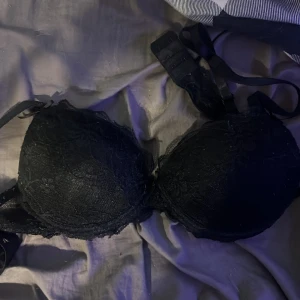 Svart spets-bh med rosett - Elegant svart bh i spets med en söt rosett framtill. Den har justerbara axelband och en klassisk design som passar perfekt under alla typer av kläder. Aldrig använd då den är för liten! 