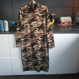 Camouflage klänning från Gina Tricot - Snygg långärmad klänning i camouflage från Gina Tricot. Perfekt för en cool och avslappnad stil. Klänningen har en hög krage och är tillverkad i ett mjukt material som ger en bekväm passform.