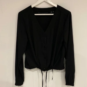 Svart blus från lager 157 - Snygg svart blus från lager 157 med v-ringning och knappar framtill. Den har långa ärmar och en justerbar knytning i nederkanten för en perfekt passform.