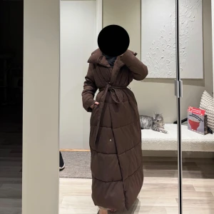 Brun lång dunjacka med bälte - Säljer en brun lång dunjacka med bälte i midjan. Jackan har en quiltad design och långa ärmar, perfekt för kalla dagar. Den har en stilren look och är både varm och bekväm. Ordpris 2000kr, använt endast 2-3 gånger 