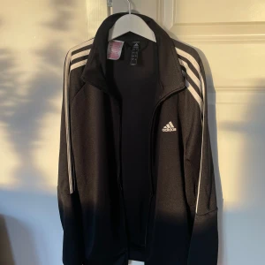 Svart träningsjacka från Adidas - Snygg svart träningsjacka från Adidas med klassiska vita ränder längs ärmarna. Jackan har dragkedja framtill och en hög krage. Perfekt för sport och fritid. Jackan är i barn storlek L och är för 13-14 åringar utifrån det som står på loggan men passar även för storlek S. Skriv privat för fler bilder och frågor🥰köpt för 500kr och mitt pris är 150kr. Priset kan även diskuteras vid snabbaffär