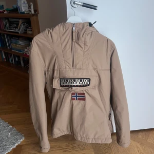 Beige jacka från Napapijri - Säljer en knapppt använd, beige jacka från Napapijri med huva och dragkedja framtill. Jackan har en stor ficka med logotyp och norsk flagga på bröstet. En bra skidjacka! 