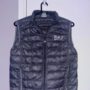 Emporio Armani väst - Snygg grå quiltad dunväst från EA7 med dragkedja framtill. Perfekt för lager-på-lager under kyliga dagar. Västen har en stilren design med EA7-logga på bröstet. Fint skick köpt för 1900 på Armanis hemsida