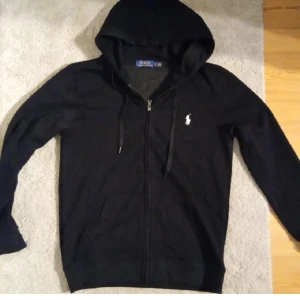 Svart hoodie från Ralph Lauren - Snygg svart hoodie från Ralph Lauren med dragkedja och klassisk logga på bröstet. Perfekt för en avslappnad stil. Den har en justerbar huva och långa ärmar.