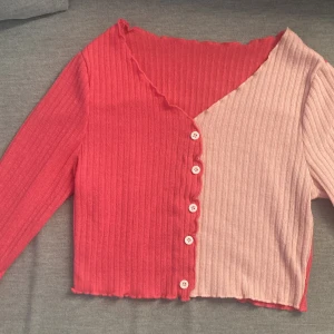 Tvåfärgad ribbad kofta från Shein - Säljer en cool tvåfärgad kofta från Shein i rött och rosa. Koftan har en ribbad struktur och knappar framtill. Den är långärmad och har en croppad design med vågiga kanter.