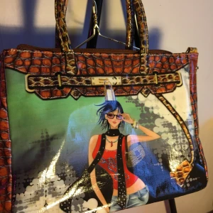 Färgglad handväska med motiv - Unik handväska med ett färgglatt motiv. Väskan har ett krokodilmönstrat brunt material och två handtag samt ett medföljande axelband.  Längd: 29cm Bredd: 16cm Höjd: 22,5cm