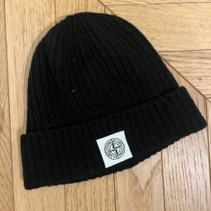 Svart stickad mössa från Stone Island - Snygg svart stickad mössa från Stone Island med ribbad design och ett broderat märke framtill. Perfekt för kyliga dagar.