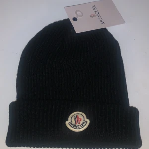 Svart mössa från Moncler - Snygg svart mössa från Moncler med ribbad design och det klassiska Moncler-emblemet framtill. Perfekt för att hålla värmen under kyliga dagar.