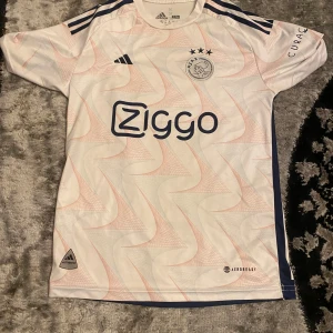 Vit Ajax fotbollströja med Ziggo-logga - Snygg vit Ajax fotbollströja från Adidas med Ziggo-logga på framsidan. Tröjan har korta ärmar och tre stjärnor ovanför klubbmärket. Den har även mörkblå detaljer och ett subtilt mönster över hela tyget. Storlek S