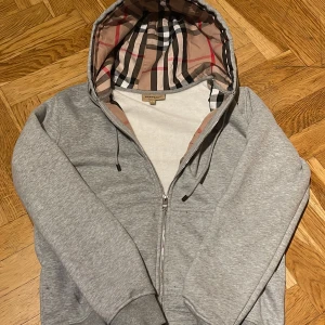 Grå huvtröja från Burberry - Säljer en stilren grå huvtröja från Burberry med klassiskt rutigt mönster i huvan. Tröjan har dragkedja framtill och en broderad logotyp på bröstet. Perfekt för en avslappnad och trendig look.