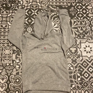 Grå hoodie från Tommy Hilfiger - Säljer en klassisk grå hoodie från Tommy Hilfiger med logotyp och texten 'Est. 1985' på bröstet. Tröjan har en justerbar huva med snören och ribbade muddar vid ärmslut och nederkant. Perfekt för en avslappnad stil.