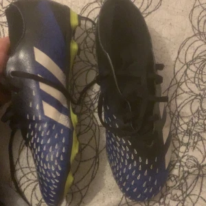 Blå och svarta fotbollsskor från Adidas - Snygga fotbollsskor från Adidas i blått och svart med vita detaljer och limegröna sulor. Perfekta för spel på gräsplaner. Skorna har snörning för en säker passform.