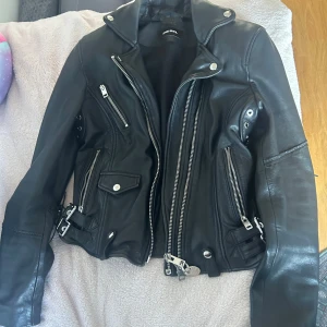 Svart skinnjacka från Diesel - Snygg svart skinnjacka från Diesel med flera dragkedjor och nitar som detaljer. Jackan har en cool bikerstil med bälten vid midjan och långa ärmar. Perfekt för en edgy look!