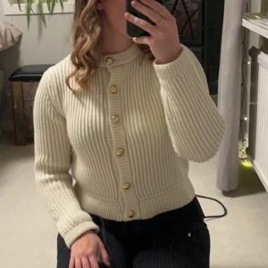 Beige kabelstickad kofta med knappar - Säljer en stilren beige ribbad kofta med guldfärgade knappar. Koftan har långa ärmar och en klassisk design som passar perfekt för kyligare dagar. Perfekt för att lägga till en touch av elegans till din outfit. Nypris ca 450kr på mango. Använd 2 gånger