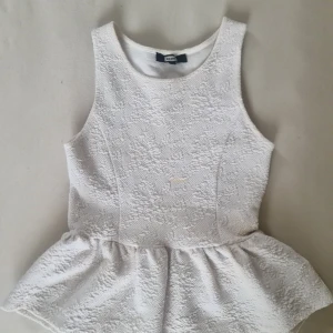 Vit ärmlös topp från Bik Bok - Säljer en vit ärmlös topp från Bik Bok i storlek XS. Toppen har en fin struktur och en peplumdetalj vid midjan. Perfekt för en stilren look.
