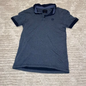 Blå pikétröja från Henri Lloyd - Snygg blå pikétröja från Henri Lloyd med broderad logga på bröstet. Tröjan har en klassisk krage och knappar framtill. Perfekt för en stilren look.