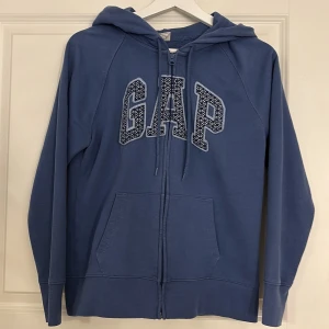 Blå hoodie från GAP - Säljer en blå hoodie från GAP med dragkedja och stor logga framtill. Tröjan har en bekväm passform med långa ärmar och en praktisk huva. Mysig inuti. Använt några gånger men är ”gått som ny” ändå. Stl S