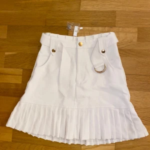Vit kjol från Amisu - Snygg vit kjol från Amisu med plisserad nederkant och dekorativa guldfärgade knappar. Kjolen har en ringdetalj och bälte i midjan för extra stil. Perfekt för en chic look!