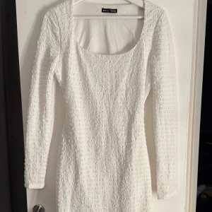 Vit klänning med öppen rygg - Elegant vit klänning med långärmad design och en stilfull öppen rygg. Klänningen har en texturerad yta som ger en unik look. Perfekt för speciella tillfällen.