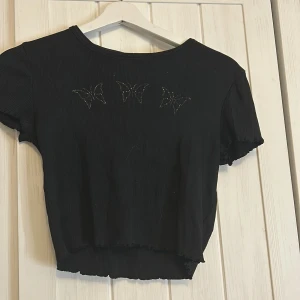 Svart ribbad topp från H&M - Säljer en svart ribbad topp från H&M med korta ärmar och ett diskret fjärilsmönster framtill. Perfekt för en stilren look. Kan även användas utomlands🤍