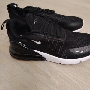 Svarta Nike Air Max 270 - Snygga svarta Nike Air Max 270 med vit och röd sula. Skorna har en modern design med synlig Air-enhet i hälen och snörning framtill. Perfekta för både stil och komfort.
