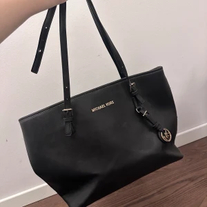 Svart axelväska från Michael Kors - Snygg svart axelväska från Michael Kors i skinn med justerbara remmar och guldfärgade detaljer. Väskan har flera fack med dragkedja för praktisk förvaring. Perfekt för att bära med sig allt man behöver. Liten defekt på ena handtaget. Därav säljer jag den för refuserad pris 
