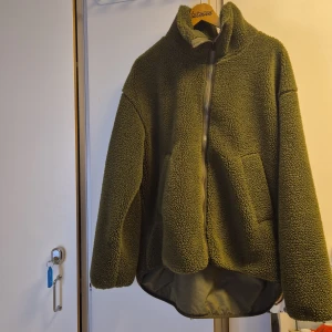 Grön teddyjacka från Stronger - Mysig grön teddyjacka från Stronger med dragkedja framtill. Jackan har en hög krage och långa ärmar, perfekt för kyliga dagar. Den har en avslappnad passform och är tillverkad i ett mjukt material som håller dig varm.
