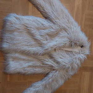 Naturvit afghan fuskpälsjackal - Säljer en fluffig beige pälsjacka i syntetmaterial. Jackan har en mysig och trendig look med långa ärmar och är perfekt för kyligare dagar. Den är tillverkad av polyester och akryl, vilket ger en mjuk och bekväm känsla.