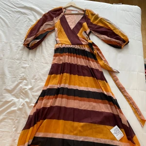 Randig omlottklänning i höstfärger - Snygg lång omlottklänning från By Malina i härliga höstfärger. Klänningen har ett randigt mönster i nyanser av gult, vinrött och rosa. Den har långa ärmar och ett knytband i midjan för en smickrande passform.