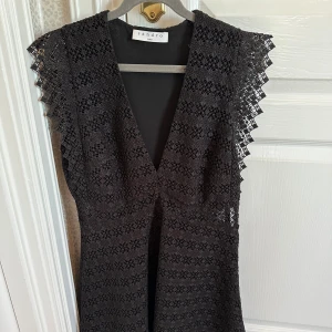 Svart spetsklänning från Sandro - Elegant svart klänning från Sandro i spets med v-ringad design. Perfekt för en stilfull look med sina detaljerade spetsmönster och ärmlösa stil.