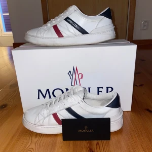 Vita sneakers från Moncler - Snygga vita sneakers från Moncler med röd och blå randdetalj på sidan. Skorna har en klassisk design med snörning och Moncler-logga på hälen. Perfekta för en stilren look. Moncler kuvert med kvitto och qr kod angår även. Nypris: 4000kr