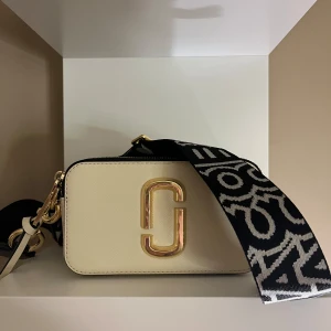 Marc Jacobs Snapshot Bag - Snygg beige axelväska från Marc Jacobs med ett stort guldfärgat emblem på framsidan. Väskan har en bred, mönstrad axelrem i svart och vitt samt dragkedja upptill. Emblemet i mitten är LITE defekt då jag råkat tappa den på marken vid ett tillfälle. Annars i väldigt snyggt skick. Nypris: mellan 3700-4000kr