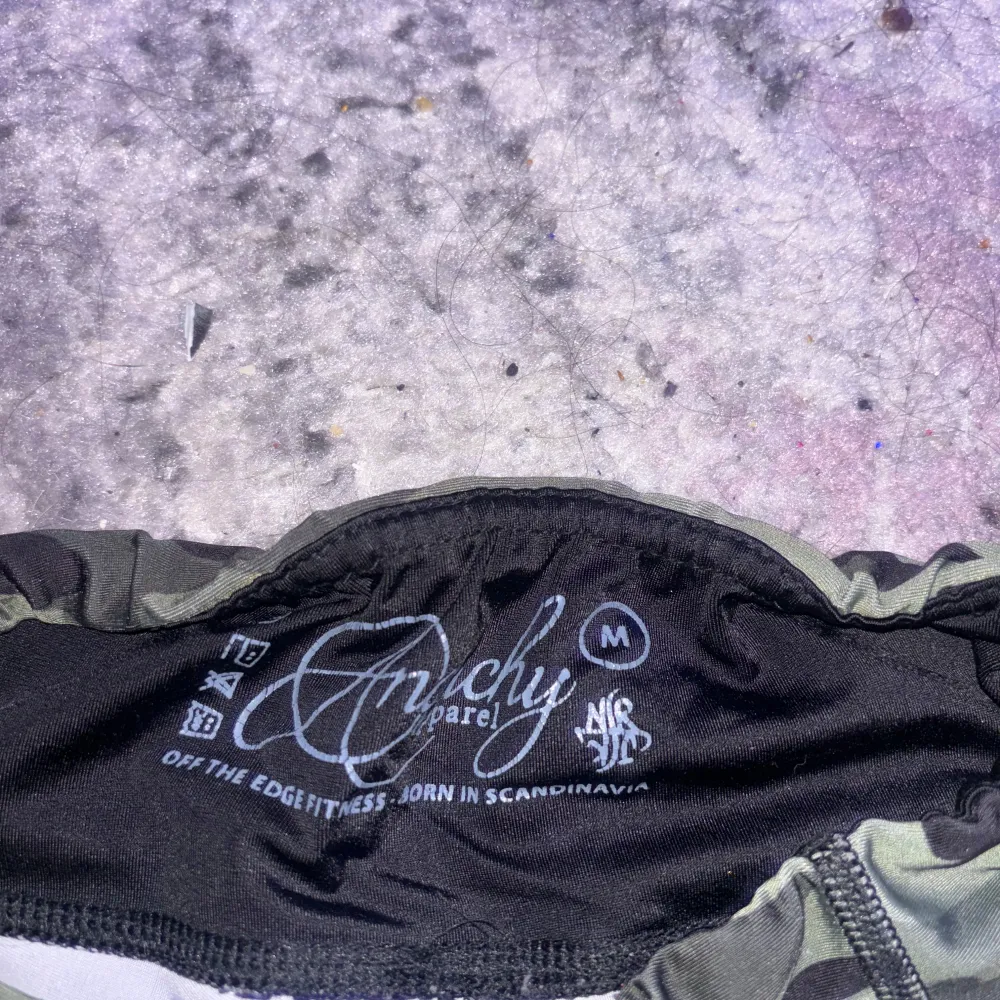 Säljer ett par coola kamouflagefärgade shorts från Anarchy Apparel. De är perfekta för träning och har en tight passform. Shortsen har en grön och svart camo-design med texten 'Whiskey Tango Foxtrot' tryckt på sidan. De är gjorda i ett bekvämt och stretchigt material, perfekt för att röra sig fritt i. Passar perfekt för sommarens alla träningspass!. Shorts.